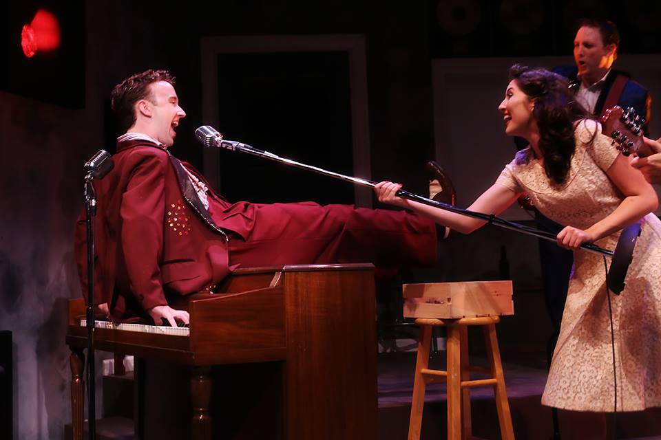 A man plays a piano while sitting on top of it. He sings into microphone stand a woman holds out to him.