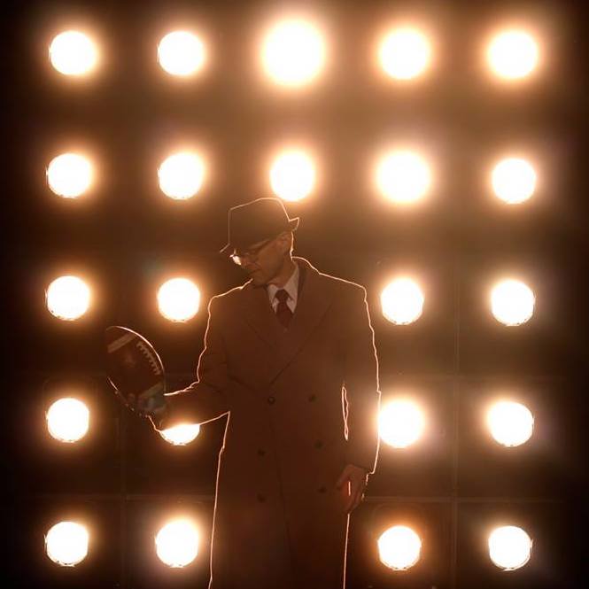 A man in a brown suit holds and stares at a football, backlit by spotlights