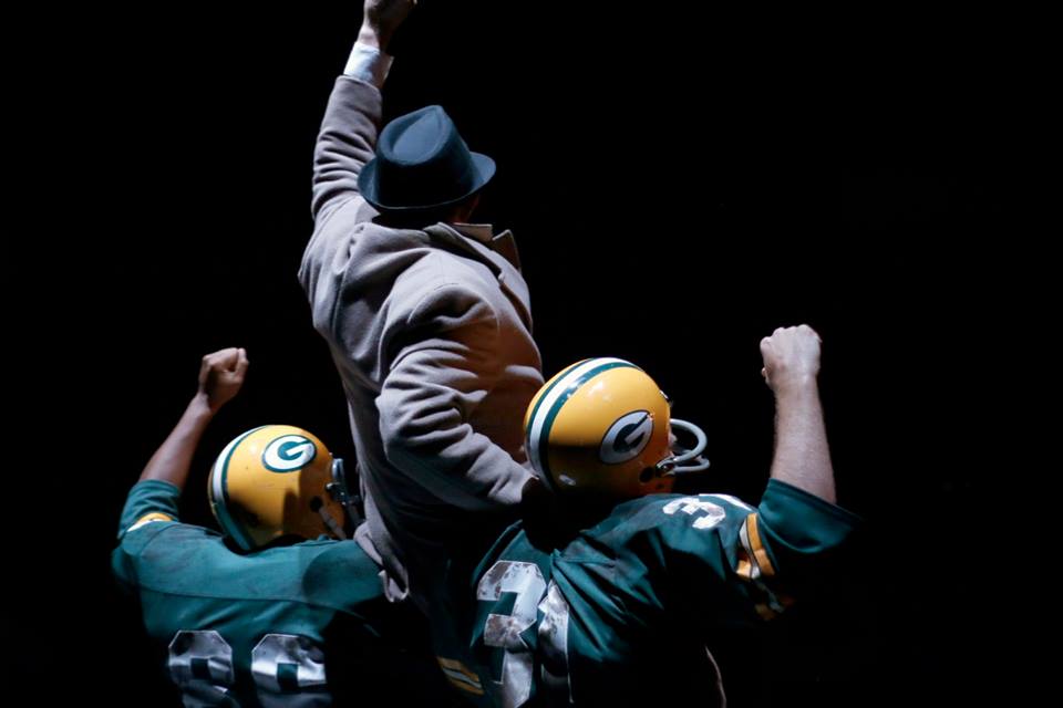 Two men in Green Bay Packers uniforms hold a man in a brown suit on their shoulders.