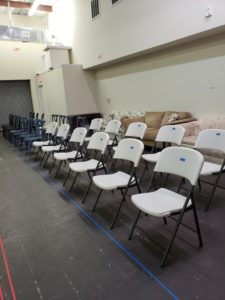 A group of 25 chairs pushed against one wall. Some are white folding chairs, other chairs are blue
