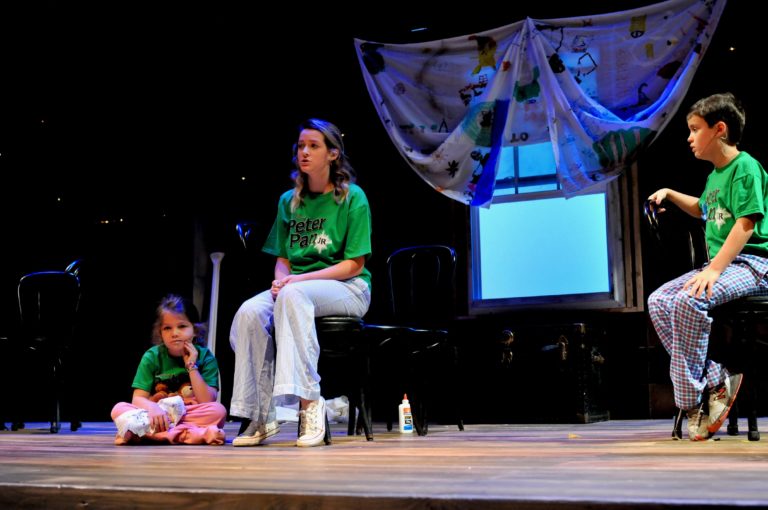 A teenage girl playing Wendy is seated center and singing while a young boy and young girl look on