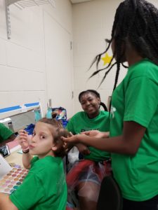 Three girls in green shirts help each other get ready in the dressing room.
