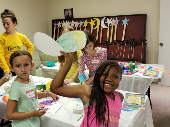 Five kids create crafts around a craft table. A dark-skinned girl holds up her creation while smiling