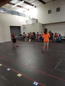 Two kids stand in the center of rehearsal space while many other kids seated in chairs watch