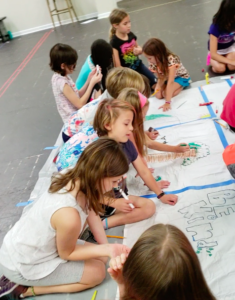A group of children kneeling on the floor, drawing on a large section of white paper