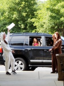 Two counselors wearing white and brown hoodie pajamas are flanking an SUV while a camper smiles out the window